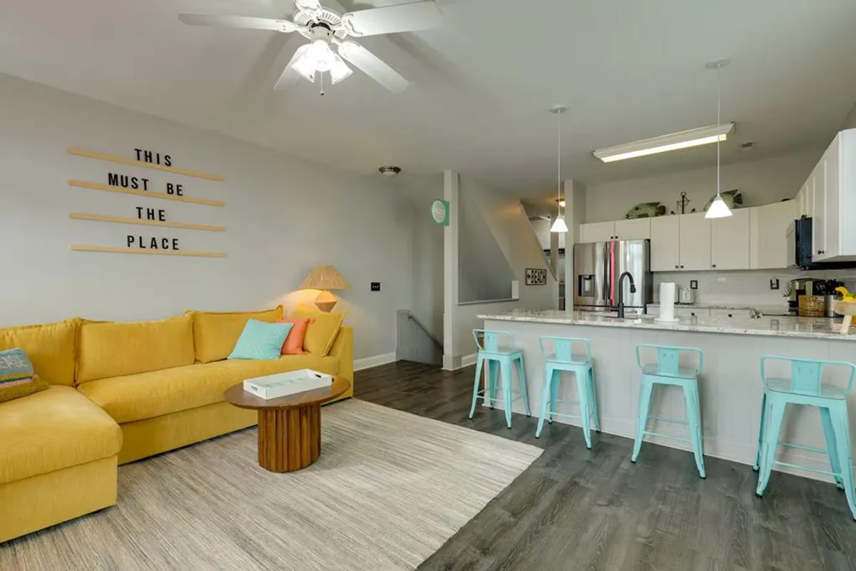 This image showcases a bright yellow sofa, bar stools at a kitchen island, and text on the wall, capturing an open-plan living room and kitchen with vibrant, welcoming decor. The space is set up for socializing, cooking, and relaxing, with ample seating and a lively atmosphere.