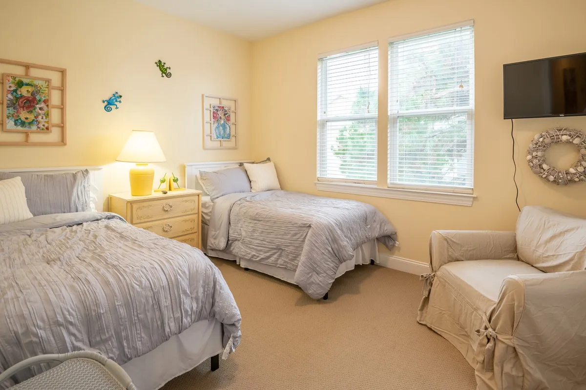 A tranquil bedroom featuring two beds with soft linens, a bedside lamp, elegant wall art, an armchair, and large windows that let in natural light. The serene atmosphere invites restful sleep and relaxation, ideal for unwinding after a day with family or friends.