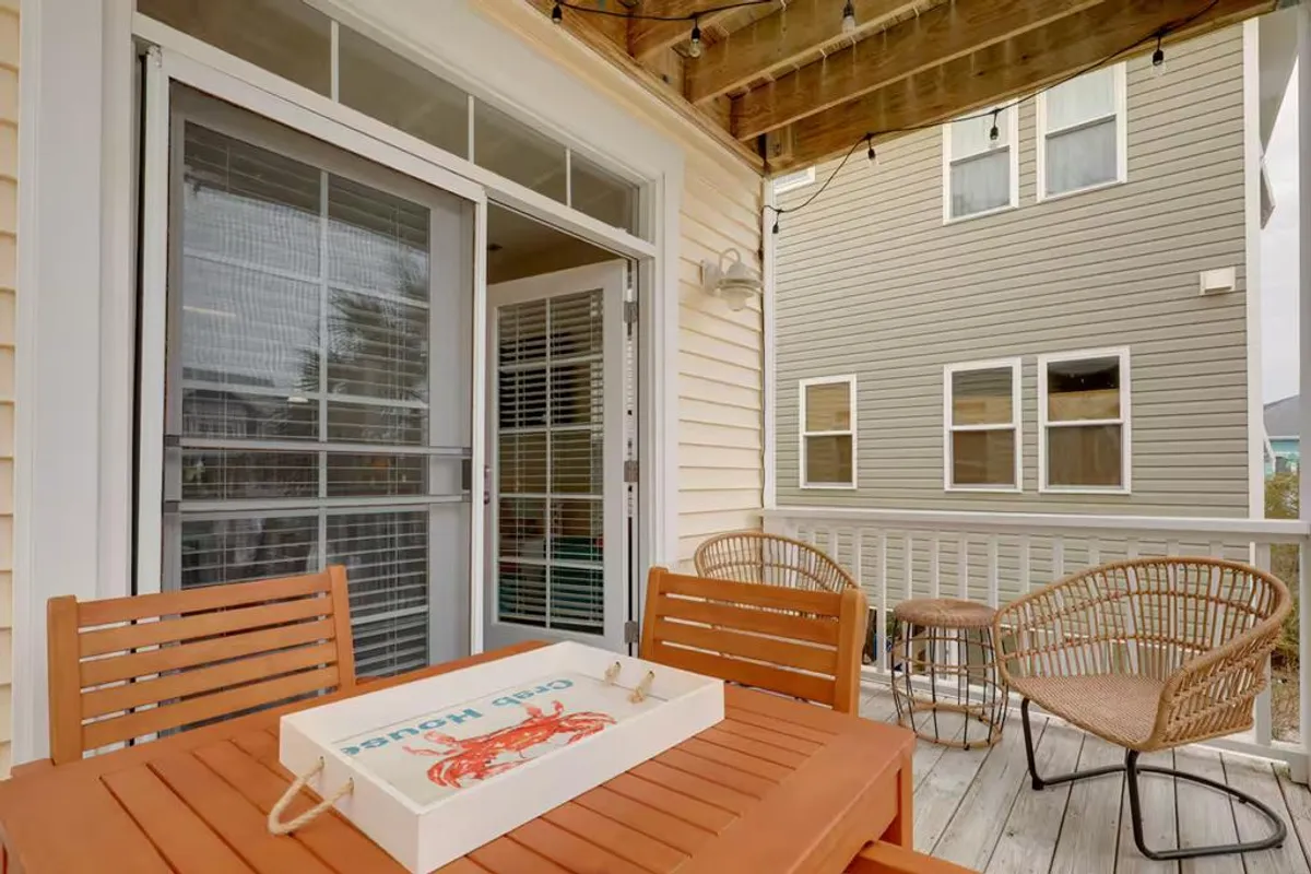A cozy exterior covered porch with a wooden table, rattan chairs, glass door, and string lights. The setting is inviting for relaxing, dining, or enjoying the ocean breeze, and visually connects the indoor social hubs to the outdoors.