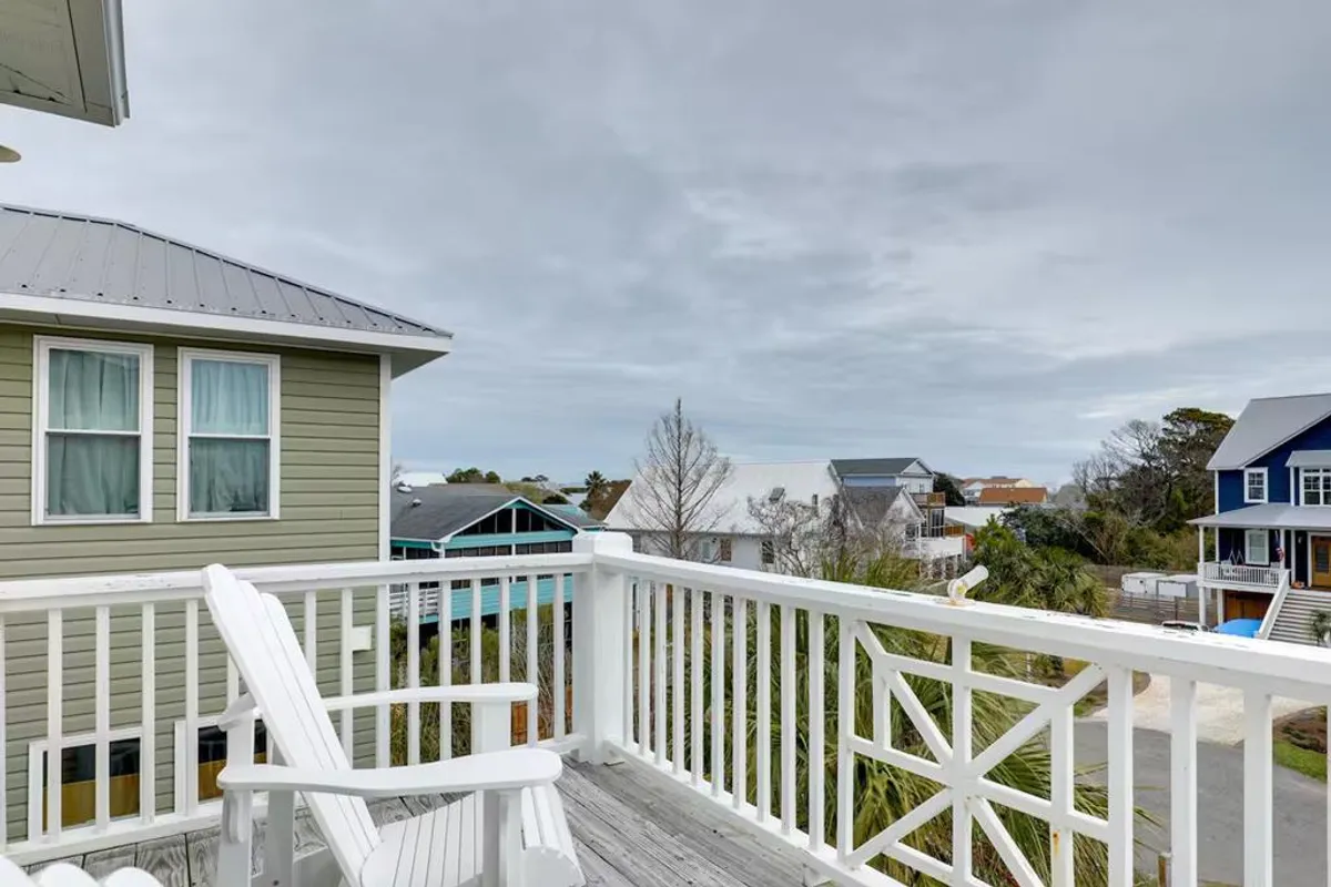 A serene exterior balcony with a white wooden chair positioned to overlook the surrounding neighborhood, bordered by a simple white railing and set against the backdrop of neighboring houses. The scene evokes a peaceful, open-air atmosphere ideal for unwinding after a day of island adventures.