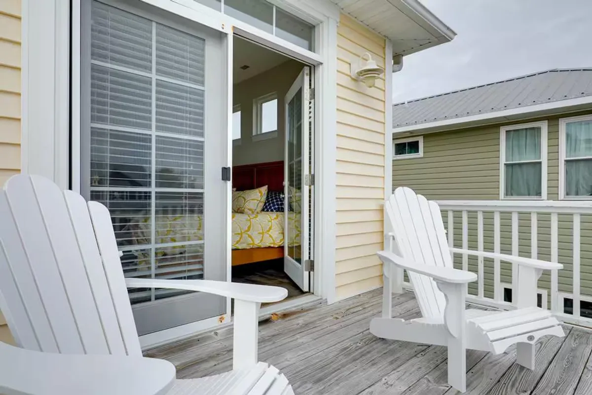 A serene deck area featuring white Adirondack chairs beside a wide open sliding door, with a glimpse of the bedroom inside. The outdoor seating faces outwards, offering a spot to enjoy the fresh air, unwind, or keep an eye on a pet enjoying the outdoors.