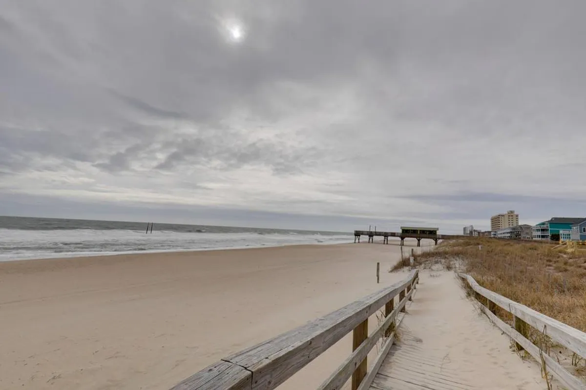 This image captures a boardwalk leading directly to the sandy beach, with ocean waves rolling in and a distant pier visible beneath a cloudy sky. The scene is tranquil and inviting, perfectly evoking the anticipation of a morning stroll to the shore.
