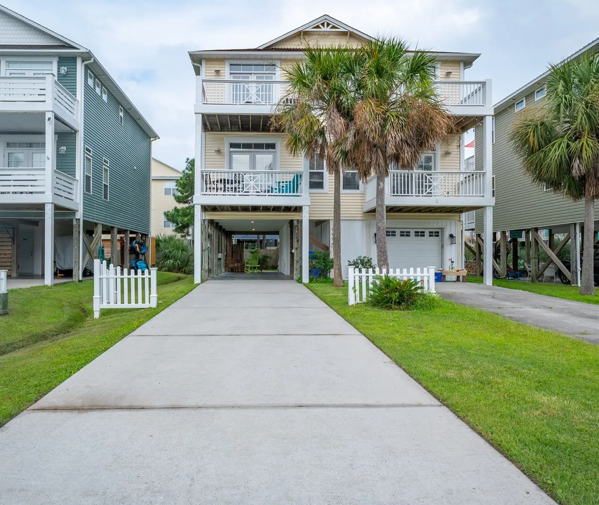 This exterior image showcases the two-story house nestled among palm trees, with a spacious driveway capable of accommodating several cars and a white picket fence enclosing a lush, grassy area. The porch and garden area are visible, suggesting spaces for family gatherings and outdoor relaxation.