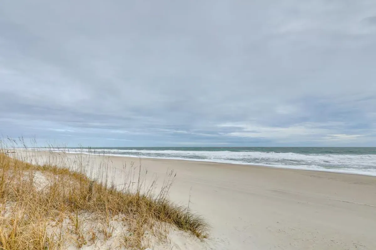 A tranquil stretch of sandy dunes meets ocean waves beneath a cloudy sky. People are seen walking along the shore, beachcombing, and snapping photos. The early morning light suggests the promise of a full day of beach adventures.