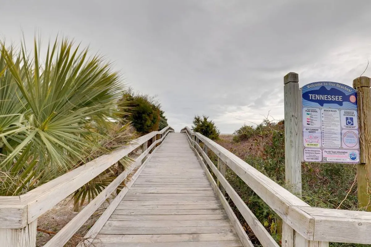 A wooden boardwalk winds through palm trees, with an informational sign hinting at local attractions or nature trails. The serene atmosphere suggests a welcoming path to exploration and beach activities.