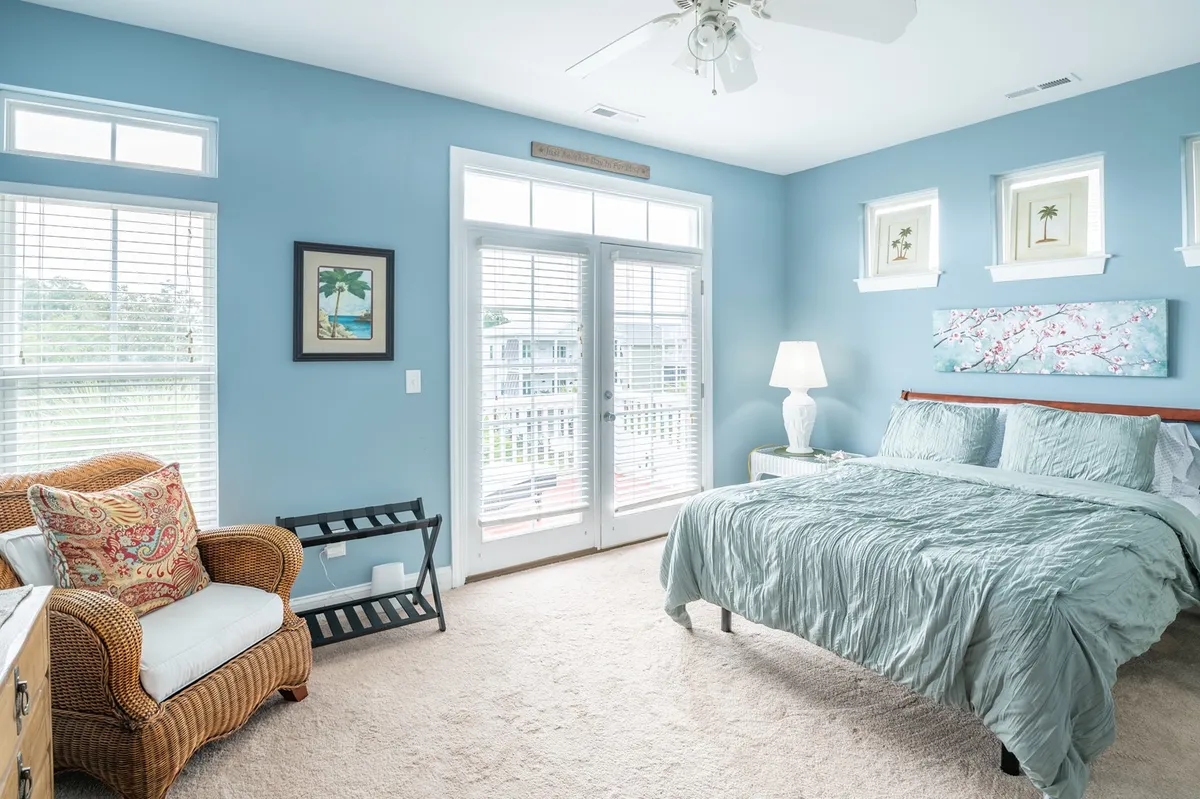 A serene bedroom with a bed dressed in light blue bedding, a comfortable armchair with a decorative pillow, double doors opening onto a private balcony, and calming artwork. The scene hints at morning coffee moments or tranquil reading with fresh air and nature just steps away.