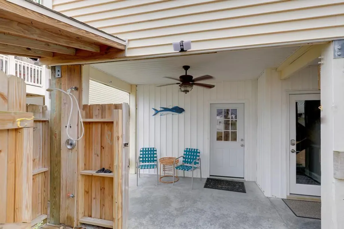 This exterior-facing view captures the front of the property, featuring an outdoor shower, decorative coastal elements, and the front door itself. Two chairs and a ceiling fan create a welcoming, practical space for transitioning between the beach and indoors. The secured entry is visible, which hints at the property’s safety features.