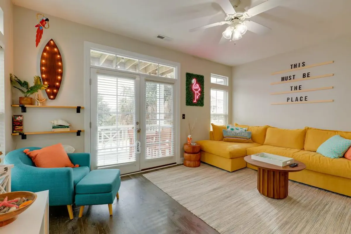 This image features a yellow sectional sofa, a neon flamingo sign, wooden coffee table, and colorful accent pillows in a cheerful living space. The setting invites guests to relax, socialize, and enjoy casual get-togethers in a comfortable, playful atmosphere.